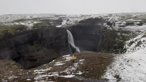 Antena:-Un-Hombre-Vestido-Con-Una-Chaqueta-Amarilla,-Caminando-Cerca-Del-Borde-De-Un-Acantilado-Hacia-La-Cascada-Granni-En-Islandia