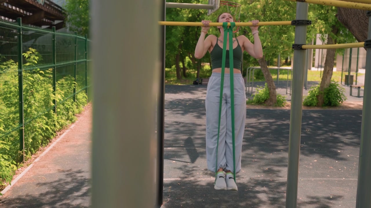 Close up of woman performing pull ups with resistance bands in outdoor park setting. Woman wears athletic clothing and uses green exercise straps attached to bar demonstrating strength
