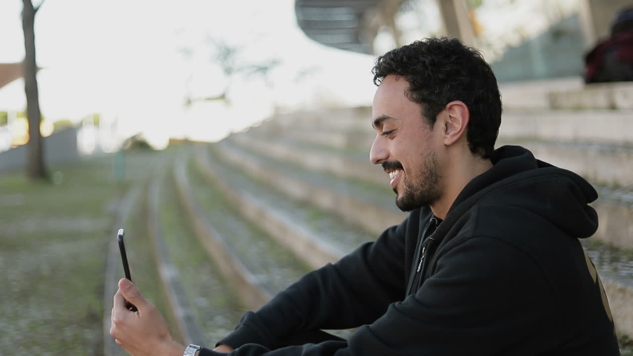 Side view of young Arabic man with dark curly hair and beard in black hoodie sitting on stairs outside