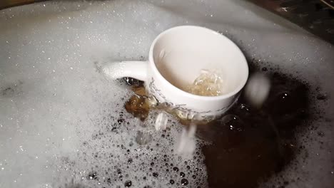 dirty dishes being washed in soapy detergent water with a tea mug falling into the sink and making a splash in slow motion