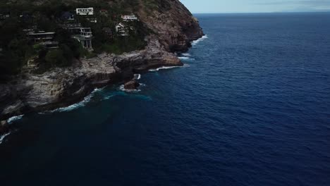 Toma-De-Un-Dron-De-Una-Playa-Junto-A-Un-Acantilado-Que-Tiene-Casas-Increíbles-Y-Bonitas-Vistas-De-Las-Olas-Rompiendo-En-Los-Acantilados-Suavizados