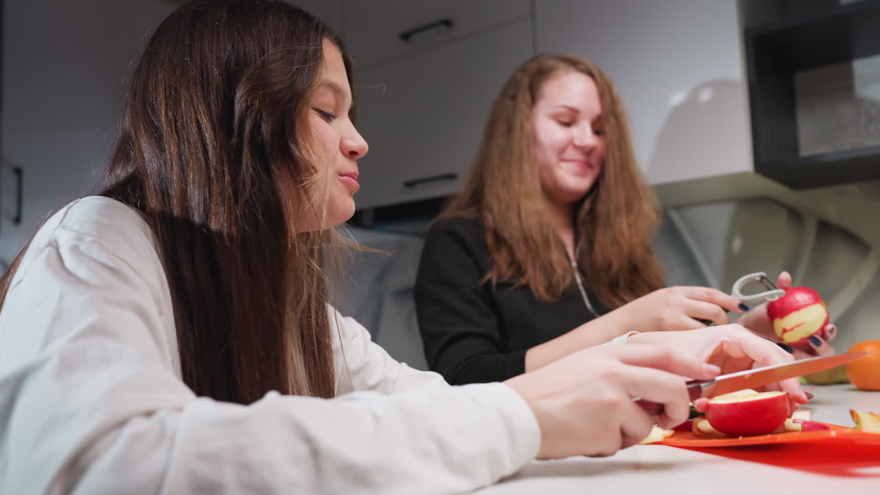 Best friends in kitchen under bright light prepare fruit; one chops apple pieces on orange board with knife, other peels skin from red apple using peeler, chatting and smiling, hands focused