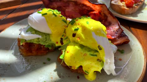 Delicious-Eggs-Benedict-breakfast-dish-with-crispy-bacon,-hollandaise-sauce-and-avocado-on-sweet-potato-hash-browns,-poached-eggs-for-brunch-at-a-restaurant,-4K-shot