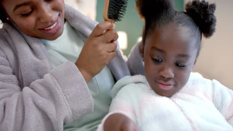 happy african american mother combing hair of daughter using tablet in bedroom, slow motion