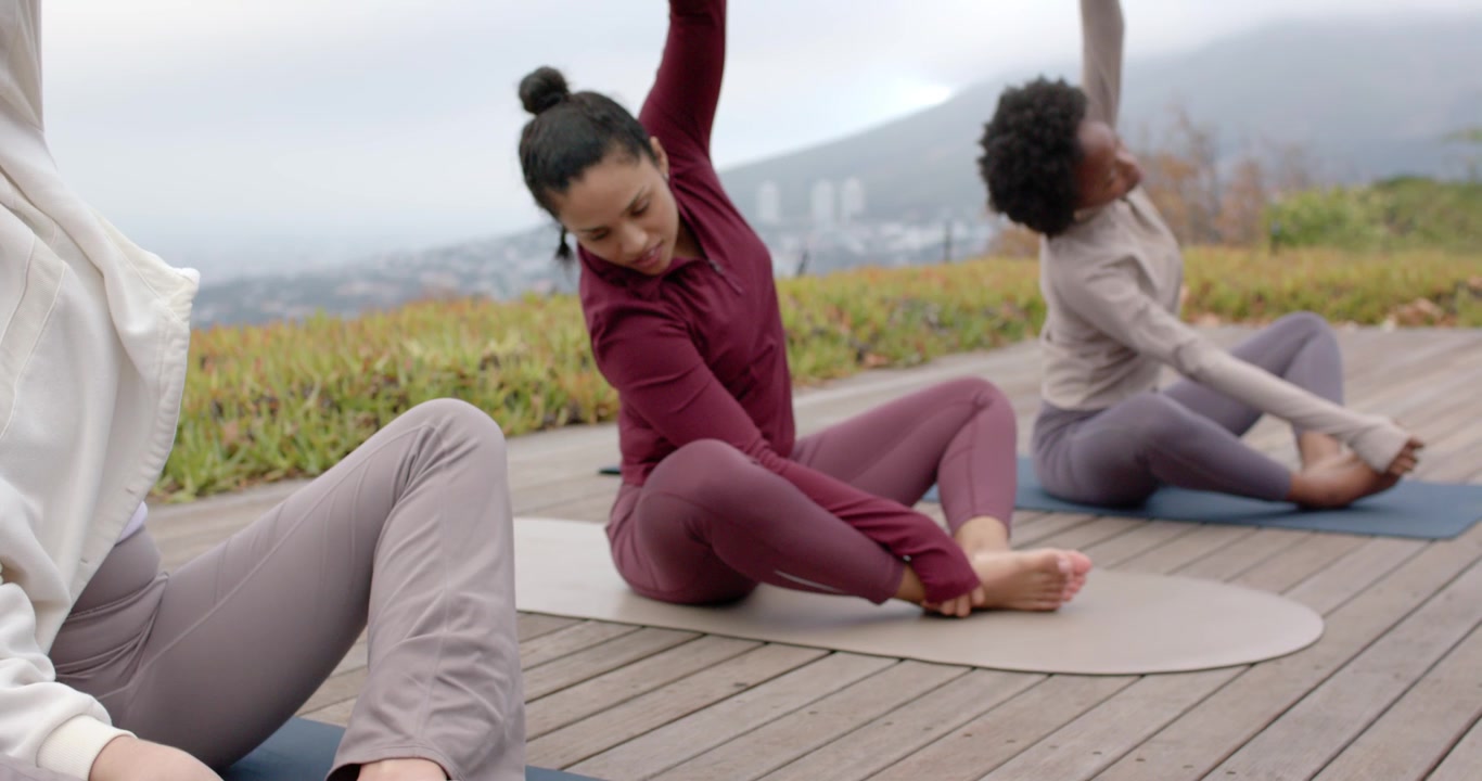 Tre donne sedute su tappetini da yoga su una terrazza, che si preparano per la pratica, facendo stretching e riscaldamento in abbigliamento sportivo.