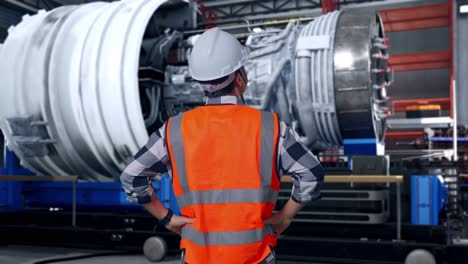 back view of a male engineer wearing safety helmet looking around while standing with arms akimbo with airplane engine maintenance conducted, analytics checking the turbine