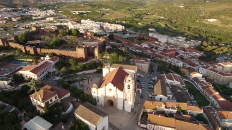 Rotierende-Antenne-Zur-Goldenen-Stunde-Zeigt-Die-Mauern-Des-Historischen-Schlosses-Sowie-Die-Alte-Kirche-In-Silves,-Portugal