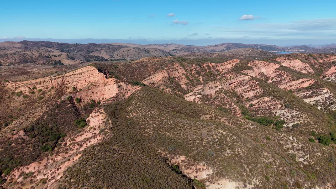 Drone footage flying above a rugged, hilly landscape with layered terrain, sparse vegetation, and a bright blue sky with scattered clouds