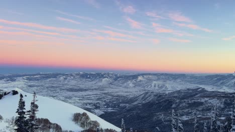 Toma-Panorámica-Hacia-La-Derecha-De-Un-Impresionante-Paisaje-Invernal-Mirando-Hacia-Un-Valle-Nublado-Cubierto-De-Nieve-Durante-Una-Puesta-De-Sol-Dorada-Desde-La-Cima-De-Una-Estación-De-Esquí-En-Las-Montañas-Rocosas-De-Utah.