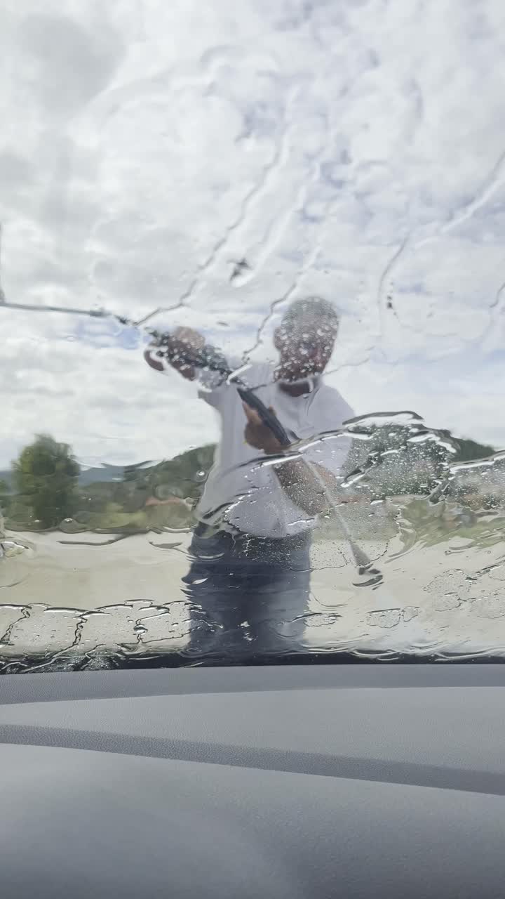 Point of view from inside a car while a man cleans the windshield with a high-pressure water jet at a self-service car wash station