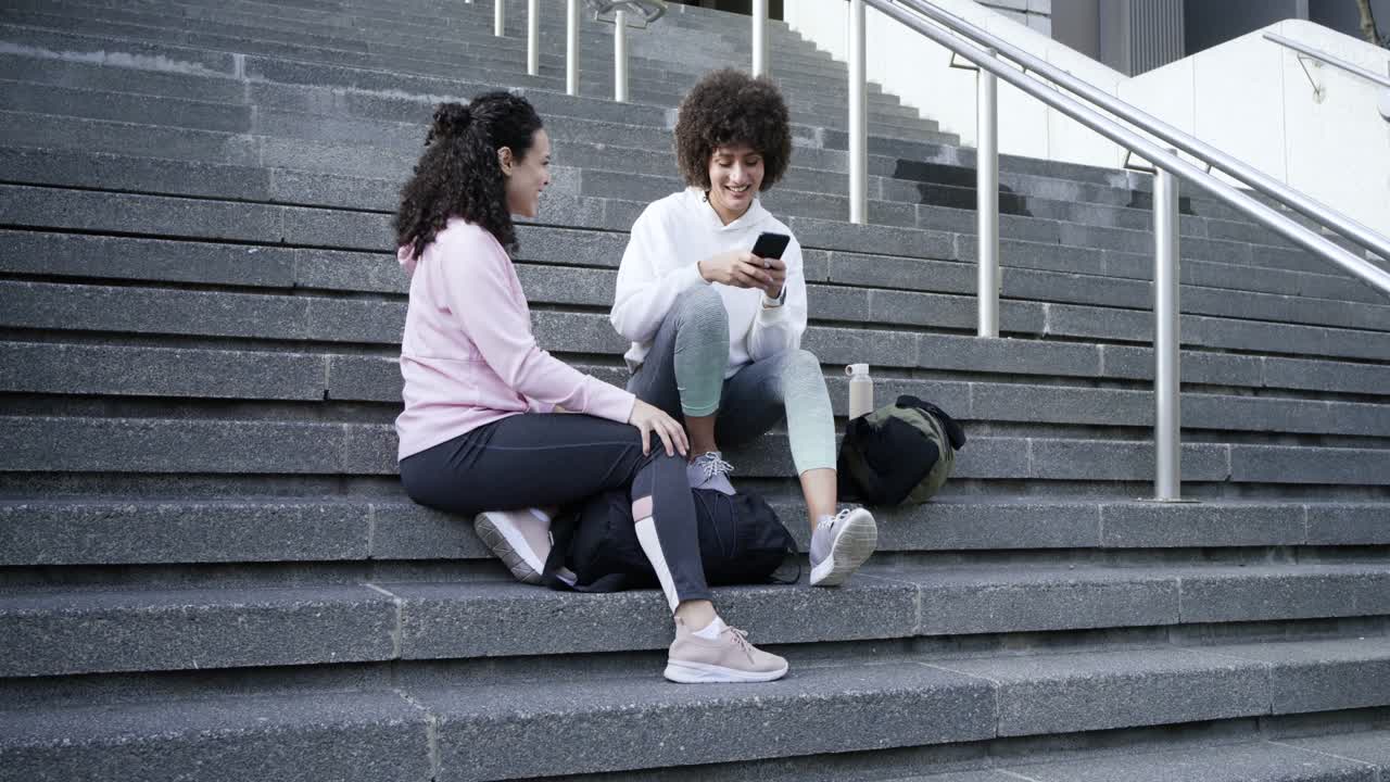 Diverse female friends scrolling smartphone content sparking chat while sitting on concrete steps