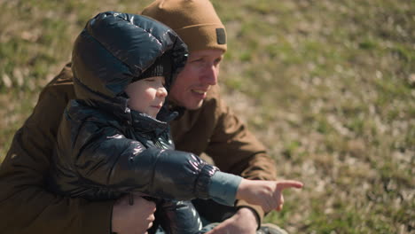 a close-up of a father squatting and carrying his son on his leg, the child, dressed in a shiny black jacket, is pointing at something and speaking with excitement