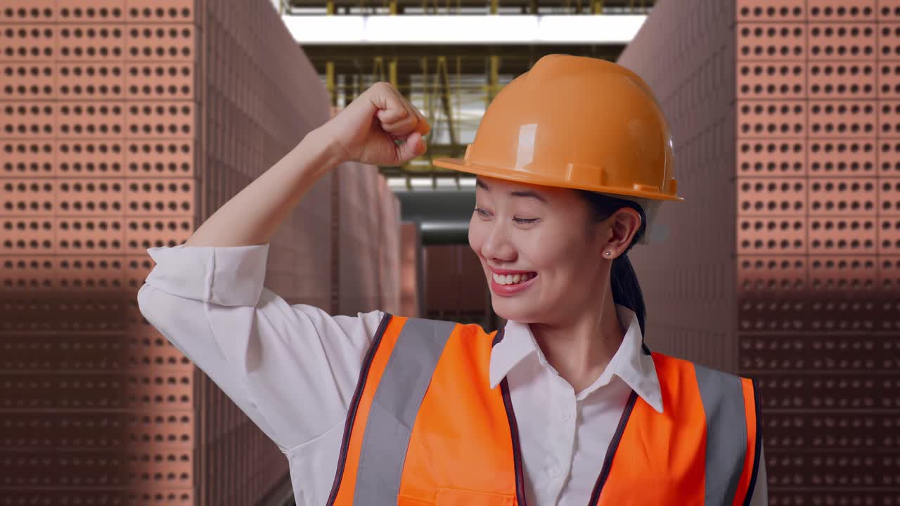Close Up Of Asian Female Engineer With Safety Helmet Flexing Her Bicep And Smiling To Camera While Standing With Red Brick Packed in Stacks Are Stored