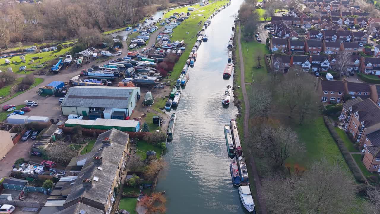Dronevlucht over de rivier Lea bij Stanstead Abbots, Engeland, schuin dalend langs de jachthaven aan de linkerkant met aangemeerde boten, terwijl huizen en bomen de rechteroever omzomen