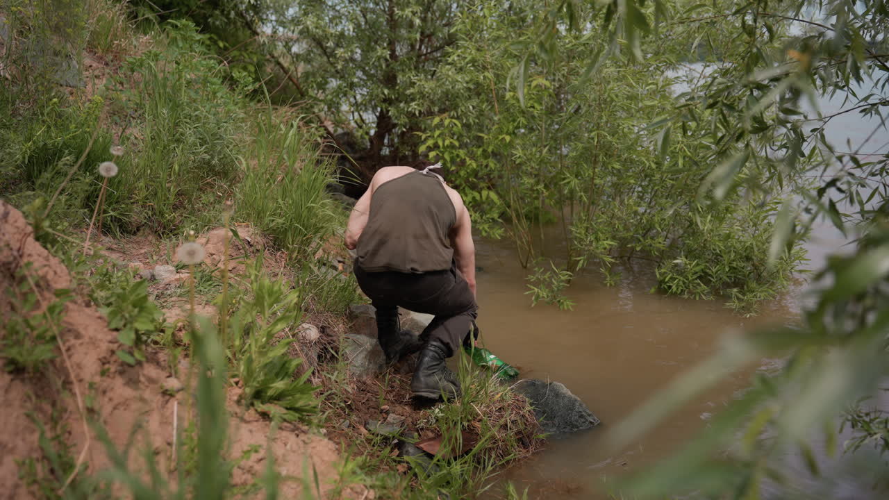 Tourist wearing tank top, gloves, and cargo pants walks down grassy riverbank holding empty bottle in hand, preparing to collect water from river surrounded by lush vegetation under cloudy daylight