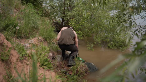 tourist wearing tank top, gloves, and cargo pants walks down grassy riverbank holding empty bottle in hand, preparing to collect water from river surrounded by lush vegetation under cloudy daylight