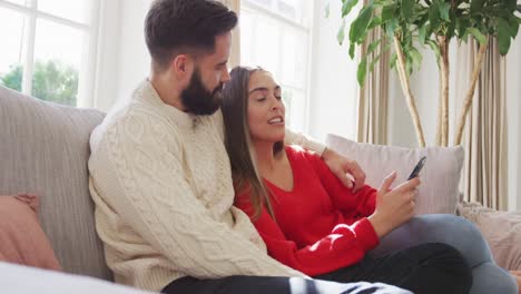 Video-De-Una-Feliz-Pareja-Caucásica-Sentada-En-Un-Sofá-Y-Usando-Un-Teléfono-Inteligente