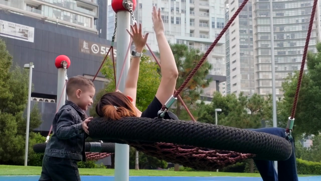 Una scena gioiosa si svolge mentre un bambino di due anni spinge giocosamente la sua giovane madre latina su una moderna altalena a letto in un vivace parco giochi cittadino. La loro interazione è piena di risate e un caldo abbraccio.