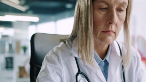 close-up view of senior caucasian female doctor sitting in hospital office working and typing on laptop at workplace