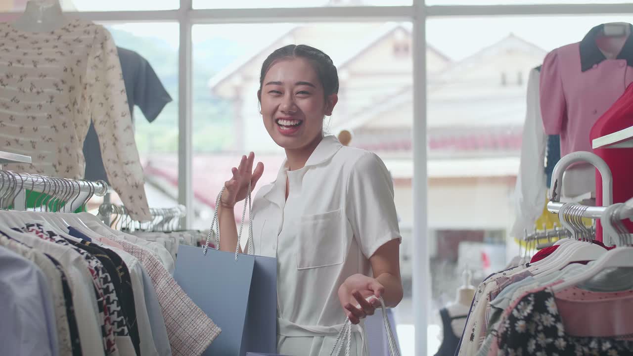 쇼핑 가방을 들고 춤추며 축하하는 즐거운 젊은 아시아 여성은 부티크에서 쇼핑하는 동안 옷을 살 수 있습니다.