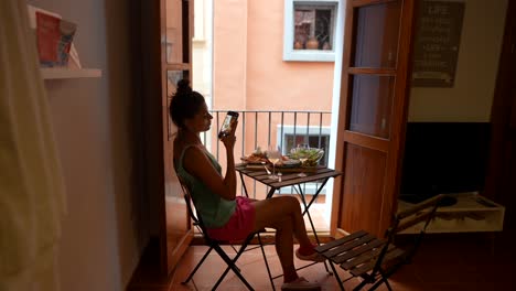 woman enjoying a meal outdoors and using mobile phone on a balcony
