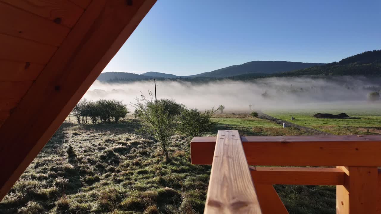 Balcony view of meadow, forest, and mountains on a frosty sunny morning. Narrow mist strips drift left, highlighting serene natural winter landscape