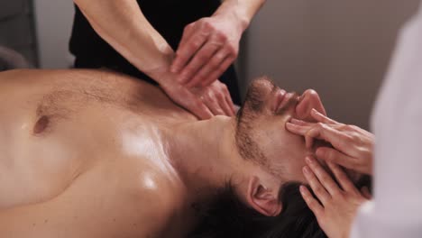 two masseuses are providing a massage to a muscular man lying on his back on a massage table in a tranquil massage room