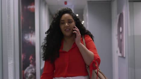 smiling biracial businesswoman talking on smartphone walking down corridor in modern office