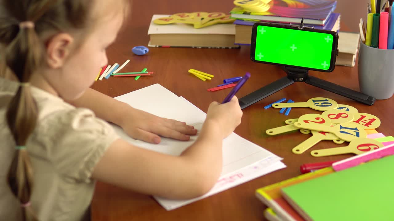 작은 여학생은 노트북에 펜을 넣고 초록색 화면의 스마트폰과 함께 테이블에 앉아 글을 쓰고 있습니다.