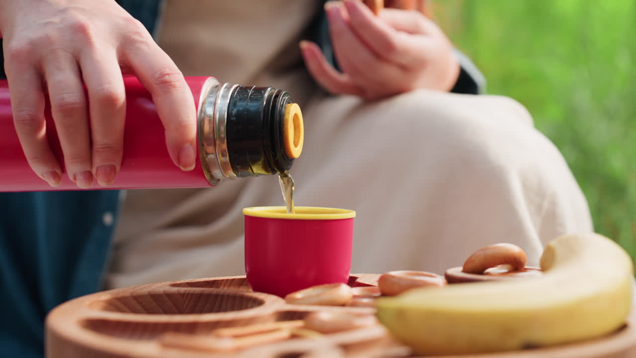 녹색 자연으로 둘러싸인 야외 여름 피크닉 설정에서 나무 피크닉 테이블에서 빨간색 보온병을 들고 바나나와 간식 옆에 있는 컵에 따뜻한 음료를 따르는 여성의 손 클로즈업