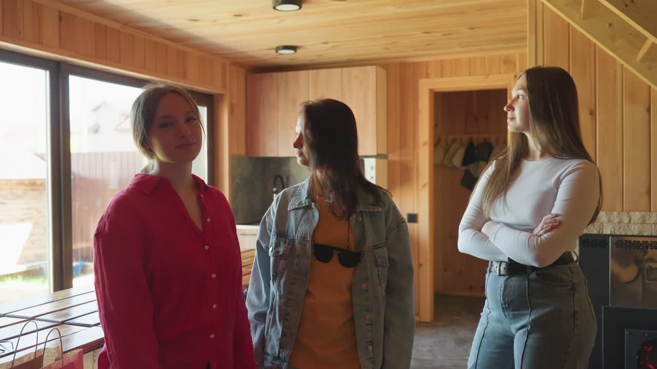 three young women stand inside wooden cabin kitchen with blank or uninterested expressions as sunlight pours in through large glass window, suggesting boredom