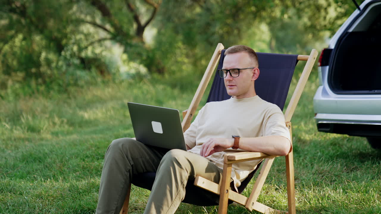 Caucasian freelancer starting work on laptop. Man opens laptop sitting in chair on the meadow. Car at backdrop.