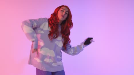 Foto-De-Estudio-De-Una-Mujer-Joven-De-La-Generación-Z-Bailando-Sobre-Un-Fondo-Rosa-Sosteniendo-Un-Teléfono-Móvil