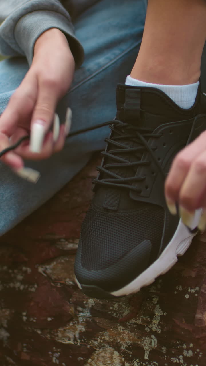 Close-up of woman with long nails tying shoelaces on black sneakers while seated on fallen tree during hike, wearing gray sweater and light blue jeans