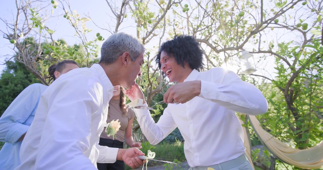 แขกหลากหลายกลุ่มกำลังเฉลิมฉลอง โดยมีชายผมหยิกเป็นผู้นำกำลังใช้มีดตัดเค้กและป้อนเค้กให้ชายอีกคนในสวน