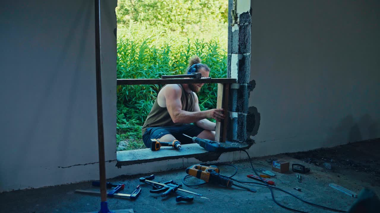 A Man is Working on a Window Frame, Using Tools to Secure a Wooden Support Into Place as Part of a Construction Project - Medium Shot