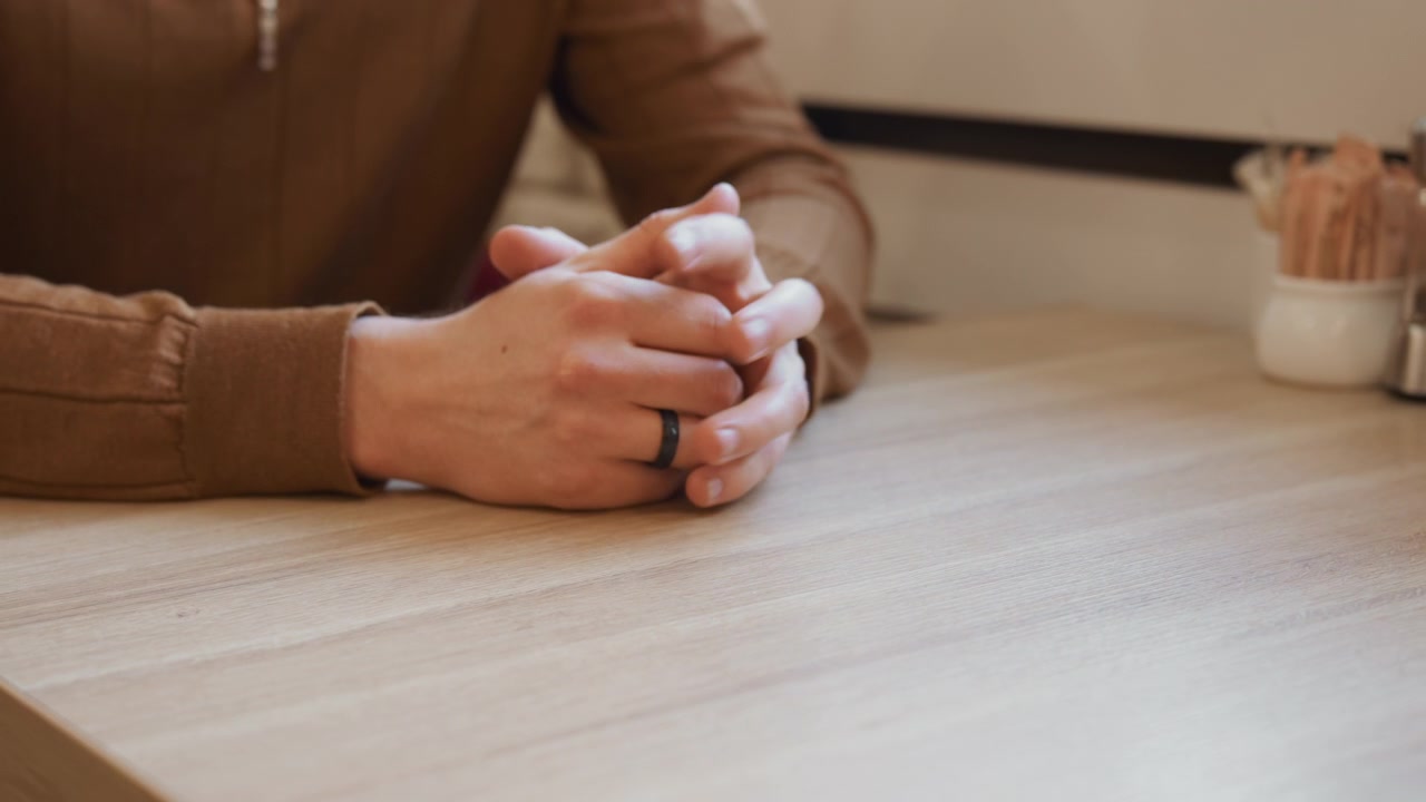 Close up of person hands resting on table fingers interlaced. subject wears brown long sleeve shirt and sits at wooden surface with soft lighting. small white container holding straws