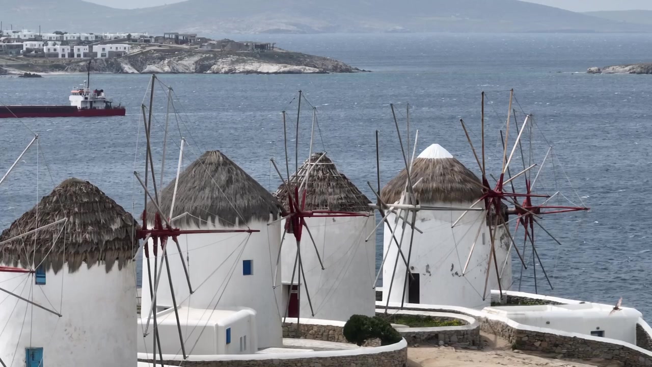 Ripresa aerea con drone dei famosi mulini a vento di Mykonos in una nuvolosa giornata invernale, che rivela una nave ancorata nel golfo protetto sullo sfondo.