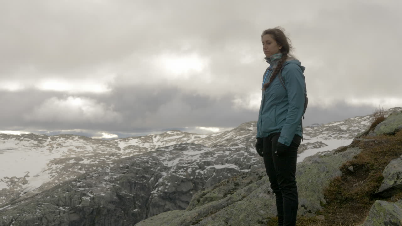 Uma mulher aprecia uma vista nevada e rochosa de uma montanha de alta altitude em um dia ventoso e nublado na Noruega, em câmera lenta