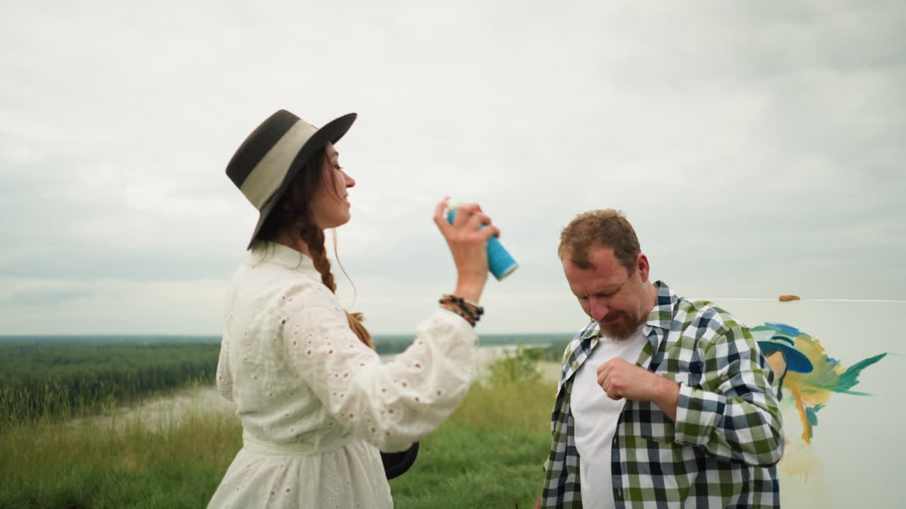 женщина в стильной шляпе и белом платье распыляет репелент от комаров на голову мужчины в пышном травяном поле. мужчина, одетый в клетчатую рубашку, спокойно стоит, в то время как женщина заботится о нем