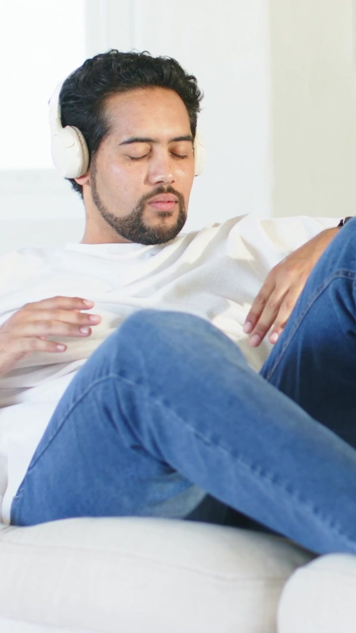 Vertical video: Listening to music, man in white tee swaying, adjusting legs on sofa with headset