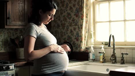 a pregnant woman gently cradles her baby bump while standing by the sink in a quaint kitchen. the warm light streams through the window, highlighting her joyful anticipation and calm demeanor.