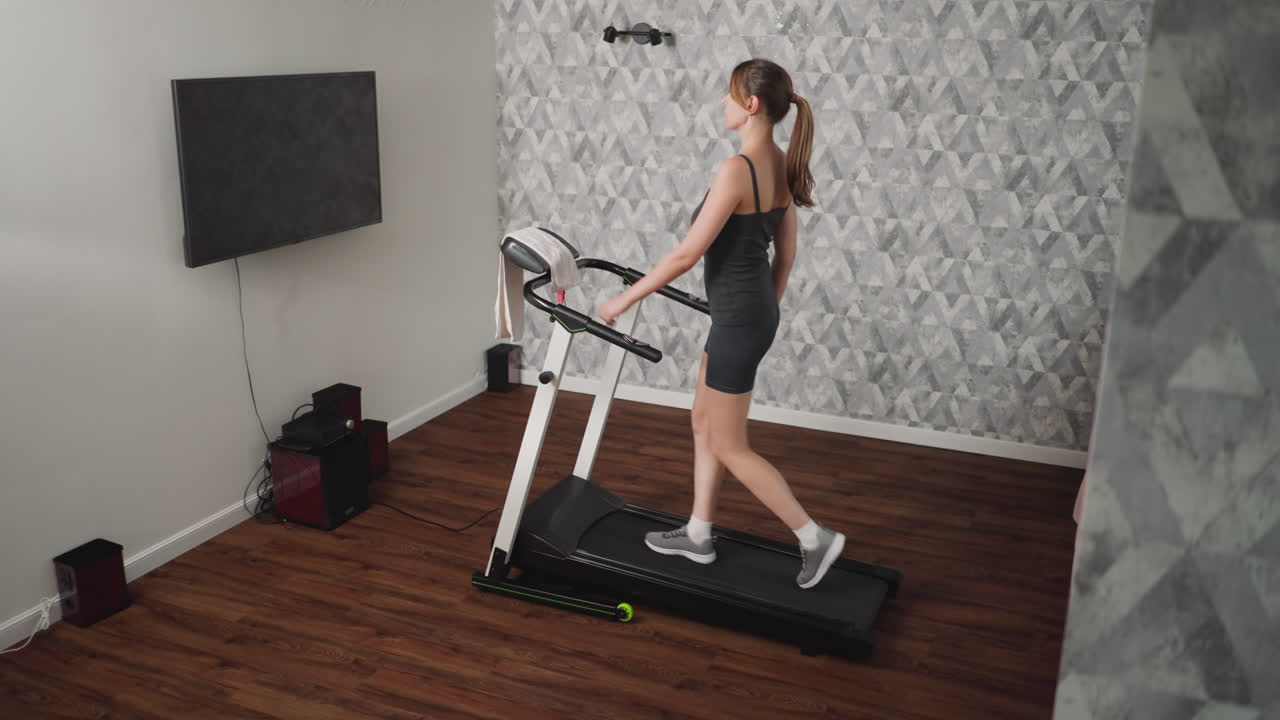 Young adult walking on treadmill during indoor fitness exercise, maintaining composed posture in athletic wear, towel placed on handlebar, patterned wallpaper and entertainment setup visible in home