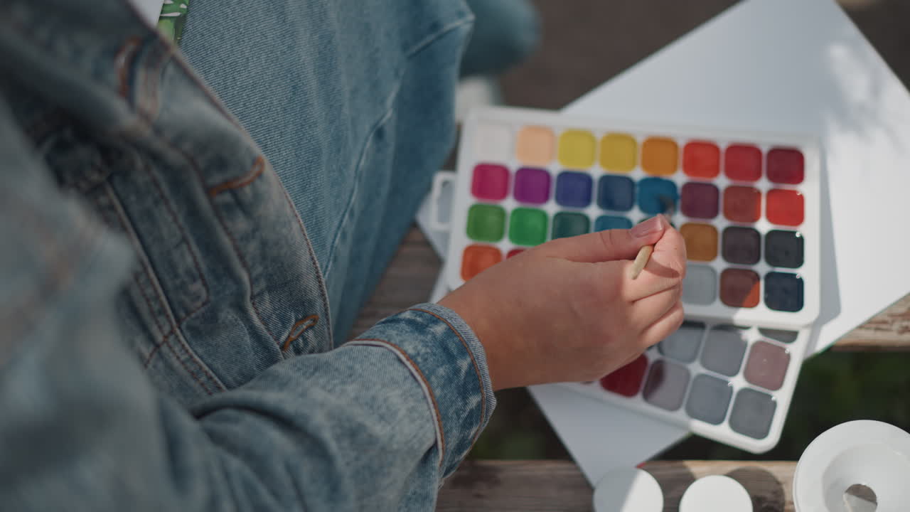 Closeup watercolor palette hand mixing tones on lap, denim jacket sleeve visible on porch, mentor tests swatches on blank paper, jars and brushes arranged, calm reflective atmosphere, experimental