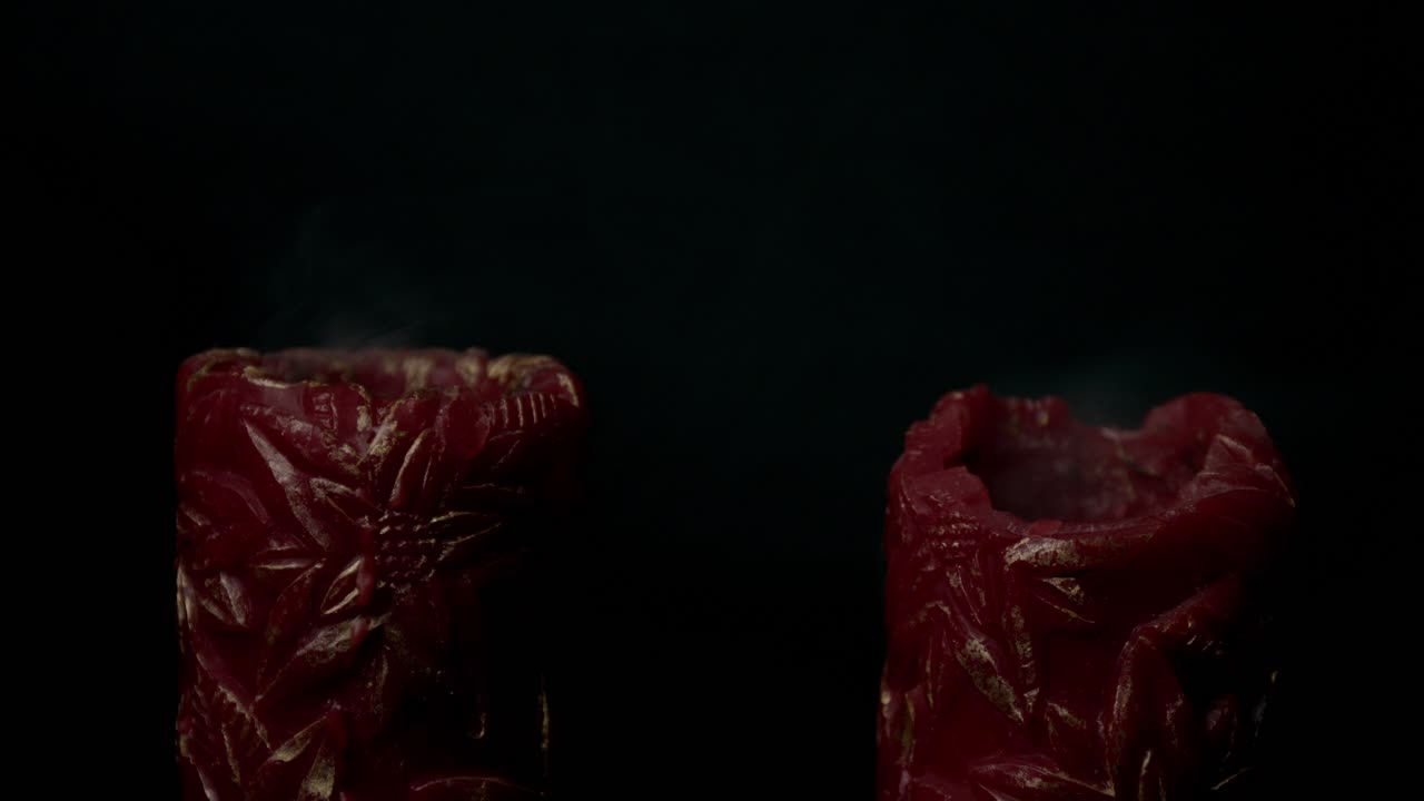 Close-up shot of two red candles with intricate designs and rising smoke against a black backdrop, symbolizing the concept of death or endings. Dramatic, moody lighting sets a somber tone
