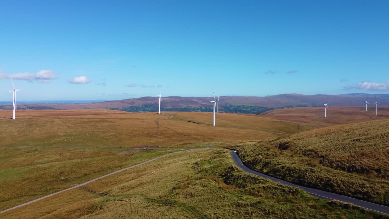 Rising Aerial Over Renewable Energy Wind Turbine Farm with Electric Car Driving Below on Rural Winding Countryside Road on Moorland Background 4K