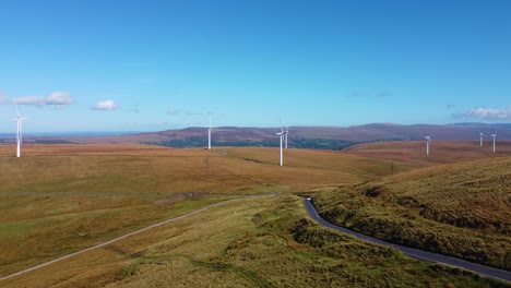 rising aerial over renewable energy wind turbine farm with electric car driving below on rural winding countryside road on moorland background 4k