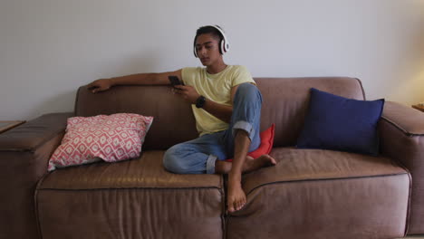 biracial man wearing headphones sitting on sofa using smartphone and smiling