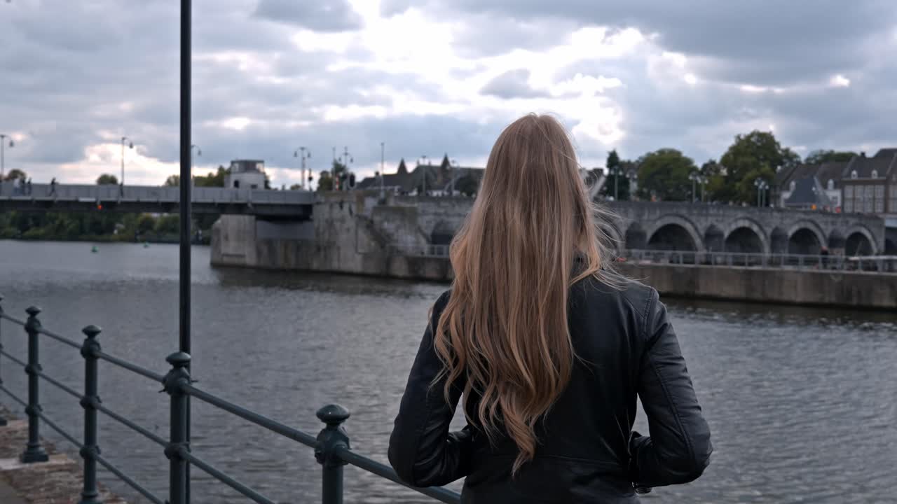 A back view of a woman with long blonde hair, dressed in a dark jacket, standing on the embankment of the Meuse River (Maas) in Maastricht, Netherlands