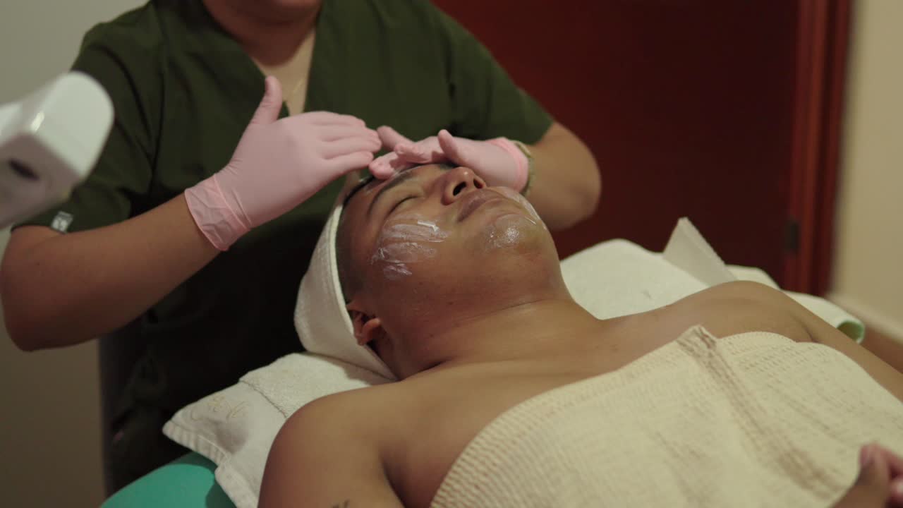 Cliente latino-americano felice che si gode un massaggio rilassante, idratante e rinfrescante e un trattamento di pulizia per la cura della pelle presso un centro benessere di un hotel resort. Uomo, applicazione di crema.